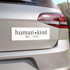 May include: A white bumper sticker with the text "human.kind (be both)" in black. The text "be both" is in rainbow colors.