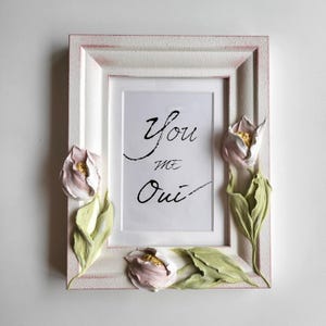 Może przedstawiać: Biała prostokątna ramka na zdjęcia z różowymi akcentami i trójwymiarowymi zdobieniami kwiatowymi. Ramka zawiera białą kartkę z odręcznym napisem "You me Oui". Kwiaty są różowe, zielone i złote.