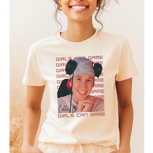 May include: A cream-coloured t-shirt with a smiling person wearing a gaming headset and cap. The shirt displays the text "GIRLS CAN GAME" and "Katie B", alongside the words "GIRLS GAME" repeated vertically.