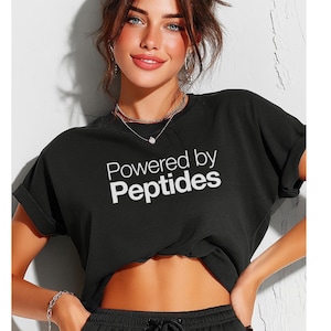 May include: Black t-shirt with the text "Powered by Peptides" in white. The shirt has rolled-up sleeves. The model is wearing a silver necklace and bracelet.