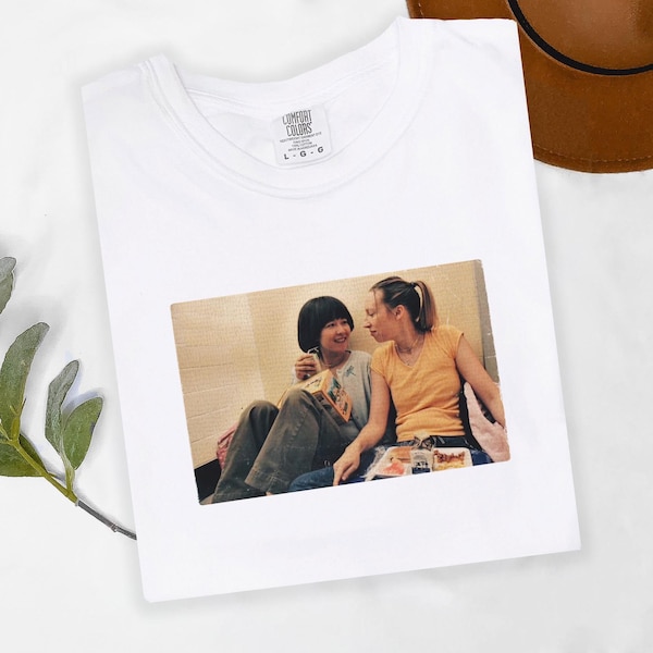 Op de afbeelding: Wit T-shirt met ronde hals met een afgedrukte afbeelding van twee personen. De afbeelding toont twee mensen in vrijetijdskleding, een met een kort zwart kapsel en de ander met een paardenstaart, zittend en naar elkaar kijkend.