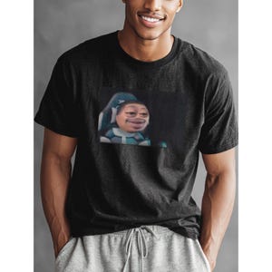 May include: Black crew neck t-shirt featuring a meme graphic. The graphic depicts a person with a distorted facial expression, seated in a gaming chair. The shirt is made of a soft material.