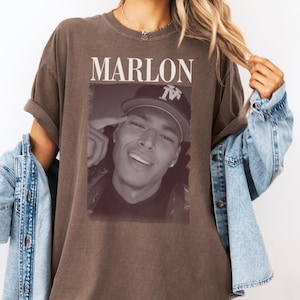 Op de afbeelding: Bruin t-shirt met de naam "MARLON" in witte letters bovenaan. Daaronder een zwart-wit afbeelding van een persoon met een baseballpet. De persoon lacht. Het t-shirt heeft opgerolde mouwen.