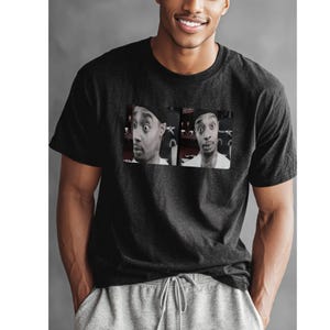 May include: Dark gray t-shirt featuring a black and white image of a person with a surprised expression. The image is split into two panels. The person is wearing a black hat and a white shirt. The t-shirt is made of a soft material.