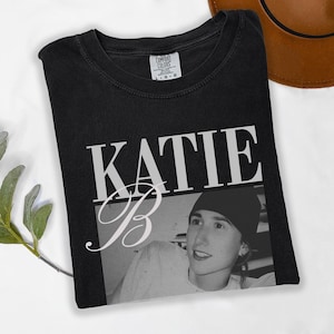 May include: Black t-shirt featuring the name "KATIE" in large white block letters above a black and white photo of a person. The letter "B" is in a cursive font. The shirt is a Comfort Colors brand.