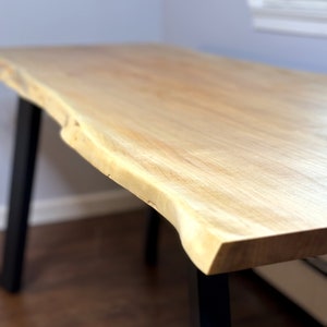 May include: A light wood desk with a live edge top and black metal legs. The desk is in a room with a white wall and a window in the background.