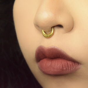 Puede incluir: Primer plano de un rostro con un aro de septum dorado. El aro tiene forma de media luna curvada. Los labios de la persona son de un color malva intenso y el tono de piel es claro. El fondo está borroso.
