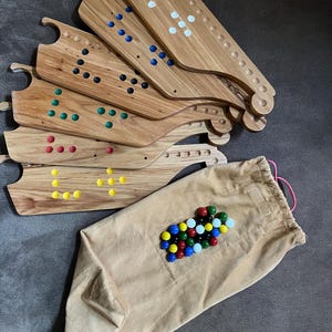 Könnte beinhalten: Eine Sammlung von Holzspielbrettern mit farbigen Punktmustern, zusammen mit einem beigefarbenen Cordbeutel, der mit bunten Murmeln gefüllt ist. Die Bretter haben einen Griff und eine Reihe von Löchern. Die Murmeln sind rot, gelb, grün, blau und weiß.
