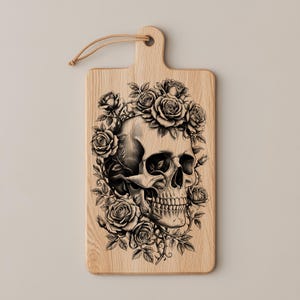 May include: Wooden cutting board with a handle and a decorative design. The design features a detailed black and white skull surrounded by roses. The board is made of light-colored wood and has a natural wood grain.