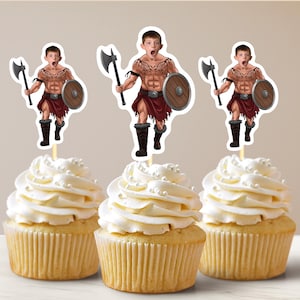 Könnte beinhalten: Drei Cupcakes mit weißem Frosting und Streuseln, verziert mit essbaren Bildern einer Person, die als Krieger mit Axt und Schild verkleidet ist.