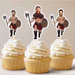 Op de afbeelding: Drie cupcakes met witte frosting en witte sprinkles, bedekt met cartoon afbeeldingen van mannen gekleed als Vikingen met bijlen en schilden.