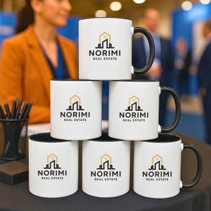 May include: Six white ceramic mugs with black interiors and handles are stacked. Each mug displays the "NORIMI REAL ESTATE" logo in gold and black. The mugs are arranged on a dark surface, with a blurred background.