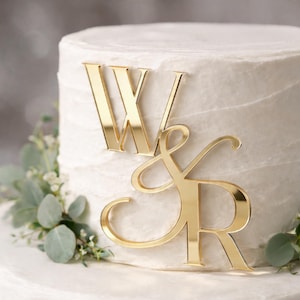 Può includere: Un topper per torta in acrilico color oro raffigurante le lettere W e R collegate da una e commerciale. Il topper è posizionato sulla parte anteriore di una torta con glassa bianca, decorata con foglie di eucalipto verdi alla base.