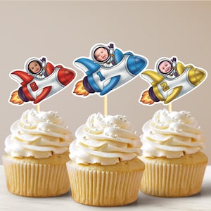 May include: Three cupcakes decorated with astronaut-themed toppers. Each topper features a child's face in a spacesuit, flying a rocket ship. The rockets are red, blue, and yellow.