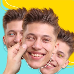 May include: A person holding a smiling face mask with three other masks in the background. The masks have different colored eyes and are set against a yellow and blue background.