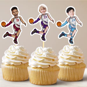 May include: Three cupcakes topped with white frosting and pearl sprinkles. Each cupcake has a basketball player cake topper. The players are wearing basketball uniforms with the number 23 and holding orange basketballs.