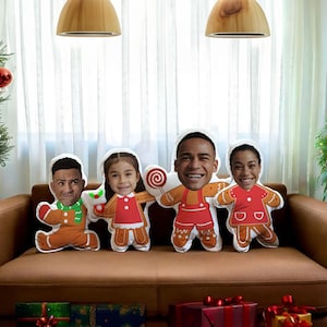 Pode incluir: Quatro almofadas em forma de homem de gengibre com rostos de pessoas em um sofá marrom. As almofadas são decoradas com glacê vermelho, verde e branco. O sofá está em frente a uma janela com cortinas brancas.