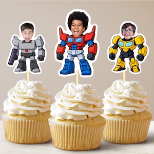 Pode incluir: Três cupcakes cobertos com glacê branco e granulado de pérolas. Cada cupcake tem um topper de personagem de desenho animado diferente com o rosto de uma criança. Os personagens são robôs em vermelho, amarelo e cinza.