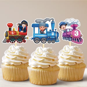 Könnte beinhalten: Drei Cupcakes, die mit Zug-Toppern verziert sind. Jeder Topper zeigt einen Cartoon-Zug mit einer Nummer und dem Gesicht eines Kindes. Die Züge sind rot, blau und lila.
