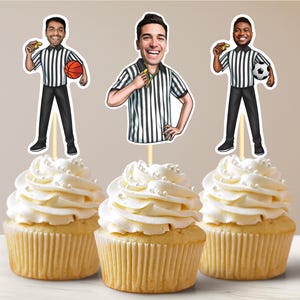 May include: Three cupcake toppers featuring cartoon illustrations of referees wearing black and white striped shirts. Each referee is holding a whistle, a basketball, and a soccer ball. The cupcakes are frosted with white frosting and white sprinkles.
