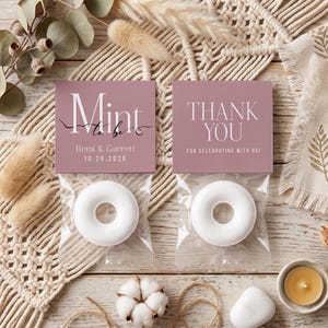 May include: Two clear plastic bags containing white ring-shaped sweets, each topped with a dusty rose card. The left card reads Mint to be, Remi & Garrett, 10.26.2026. The right card reads THANK YOU FOR CELEBRATING WITH US in white lettering.