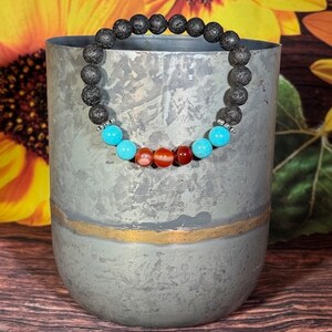 Op de afbeelding: Een zwarte lavastenen armband met turquoise en rode edelsteen kralen. De armband ligt op een grijs metalen oppervlak.