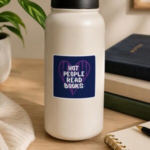 May include: A white water bottle with a black lid and a sticker that reads "HOT PEOPLE READ BOOKS" in white text inside a purple heart on a navy blue background. The bottle is on a wooden surface next to books and a notebook.