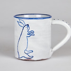 May include: White ceramic mug with a blue rim and handle. A blue outline of a rabbit is painted on the side. The handle has small blue dots. A charming and whimsical design for a coffee or tea cup.