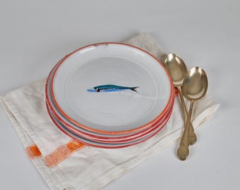 Peut inclure: Une pile d'assiettes en céramique blanche avec un motif de poisson bleu peint à la main et des bords rouges. Les assiettes reposent sur un linge blanc avec des rayures orange, accompagnées de deux cuillères en argent vintage.