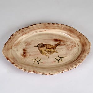 Pode incluir: Travessa oval de cerâmica com um desenho de pássaro pintado à mão. A travessa bege tem uma borda ondulada e detalhes em marrom. O pássaro é marrom e amarelo com galhos verdes embaixo.