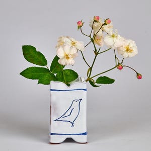以下が含まれることがあります： 青い鳥のイラストと青い縁取りが施された白いセラミック花瓶。白い花と緑の葉が入っています。花瓶は長方形で、上部はスカラップになっています。