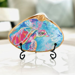 May include: A colorful sea turtle painted on a gold seashell with a black metal stand. The turtle is swimming in a blue ocean with coral reefs.