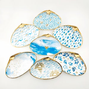 May include: Set of seven seashell trinket dishes with gold accents. Each dish features a different blue and white design, including waves, fish, dots, leaves, and a sunset.
