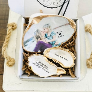 May include: A white box with a gold-trimmed oyster shell containing a photo of a mother and daughter. The shell has the text "Myla Mama" with a heart symbol. Two smaller oyster shells are in the box, one with the text "best mom ever" and the other with the text "Like the ocean, a mother's love is vast, deep, & unyielding."