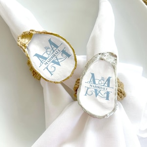 May include: Two oyster shell napkin rings with a gold and silver finish. The rings are decorated with a blue monogram and the name "MARSHALL".
