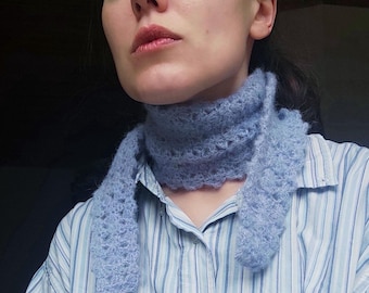 Peut inclure: Écharpe bleu clair tricotée à la main avec un motif ajouré délicat. L'écharpe est faite d'une laine douce et duveteuse et est portée autour du cou. La personne porte une chemise boutonnée à rayures bleues et blanches.