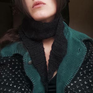 Pode incluir: Um cachecol preto de malha é usado sobre um casaco estampado verde e preto. O cachecol tem um design texturizado. O casaco tem gola e acabamento verdes, com um padrão preto e branco no corpo. O casaco está abotoado.