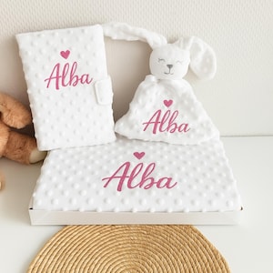 Peut inclure: Un ensemble cadeau pour bébé blanc comprenant une couverture, un petit animal en peluche et une housse, tous avec un motif texturé à pois. Le nom "Alba" est brodé en rose avec un cœur au-dessus sur chaque article. L'ensemble est présenté dans une boîte blanche.