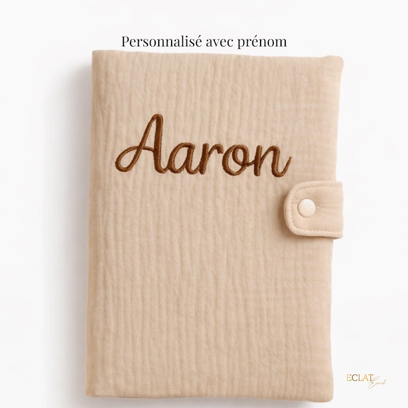 May include: A beige, textured fabric cover with the name "Aaron" embroidered in brown script. The cover has a button closure and the text "Personnalis&eacute; avec pr&eacute;nom" at the top.