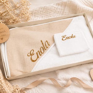 Peut inclure: Une serviette de bain à capuche pour bébé et un gant de toilette assorti dans une boîte cadeau. Les deux articles portent le prénom Enola en écriture dorée pailletée. Une étiquette en bois avec le texte Fait avec amour pour bébé est incluse.