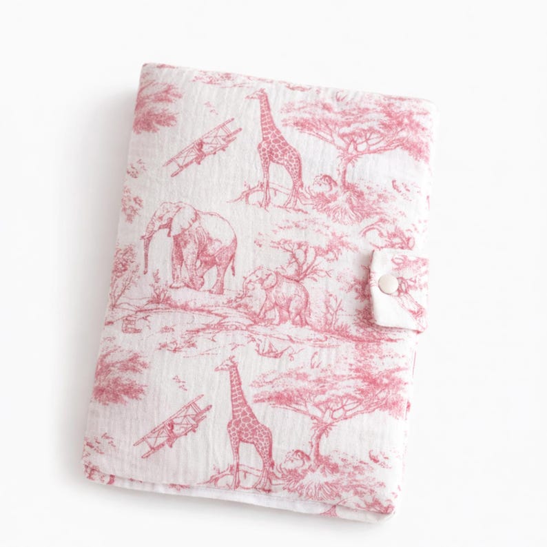 May include: A white and pink toile fabric baby blanket featuring a safari scene with giraffes, elephants, and trees. The blanket has a button closure. The fabric has a crinkled texture.