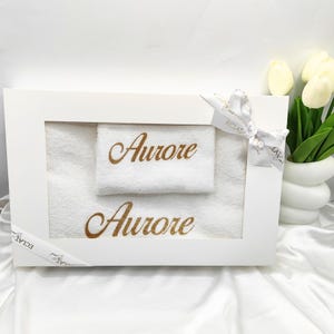 Peut inclure: Une boîte cadeau blanche avec un ruban blanc et une serviette blanche avec le nom "Aurore" brodé en paillettes dorées. La boîte est posée sur une surface blanche.