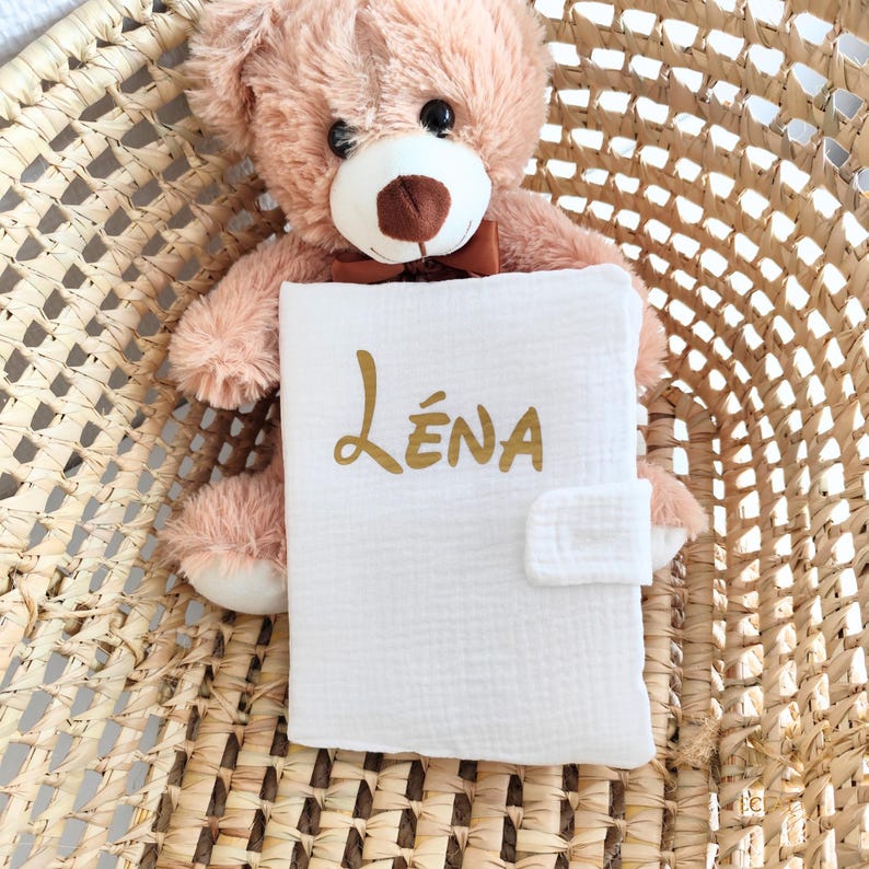 May include: A white baby book with the name "L&eacute;na" in gold script is held by a light brown teddy bear with a brown bow tie. The book has a fabric closure and is set against a woven basket.