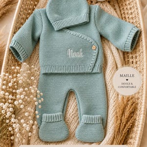 Può includere: Un set in maglia color verde salvia per neonati composto da un cardigan a portafoglio a maniche lunghe con un bottone in legno, pantaloni con piedini abbinati e un berretto. Il cardigan presenta il nome Noah ricamato. Un'etichetta circolare riporta MAILLE DOUCE & CONFORTABLE.
