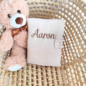 May include: A light pink baby book with the name "Aaron" embroidered in brown thread. The book is closed with a button clasp and rests in a woven basket next to a plush teddy bear.