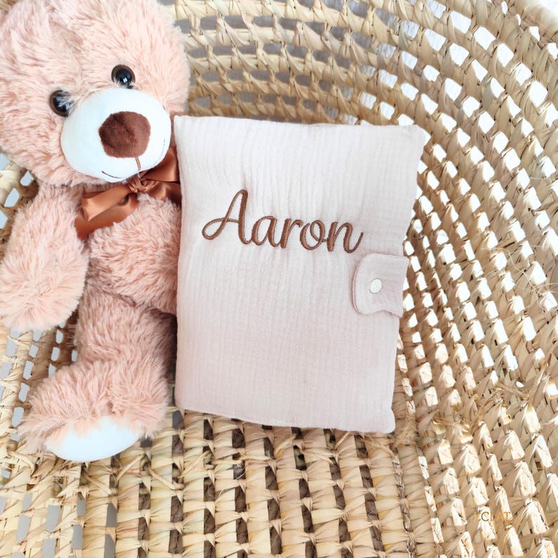 May include: A light pink baby book with the name "Aaron" embroidered in brown thread. The book is closed with a button clasp and rests in a woven basket next to a plush teddy bear.