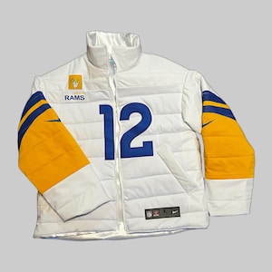 Puede incluir: Chaqueta acolchada blanca con detalles azules y dorados. El número 12 en azul en la parte delantera, con la palabra "RAMS" y un logotipo del equipo. Las mangas tienen rayas doradas y azules. Incluye cremallera completa y logotipo de Nike.