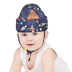 May include: A navy blue baby safety helmet with a colorful space-themed pattern, including rockets and planets. The helmet has a red trim and a chin strap. The baby is wearing a white tank top.