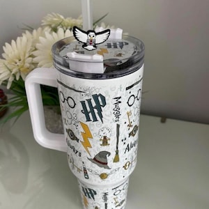 May include: A white insulated tumbler with a handle and a clear lid featuring a straw and an owl topper. The exterior is decorated with a pattern of black, gold, and grey icons including glasses, lightning bolts, a sorting hat, and the text HP and Muggles.