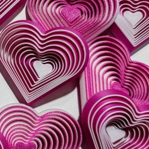 May include: Close-up of several pink and white heart-shaped objects. Each heart has a layered design, with concentric hearts decreasing in size towards the center. The hearts are arranged at different angles, creating a visually appealing pattern. The overall theme is love and romance.
